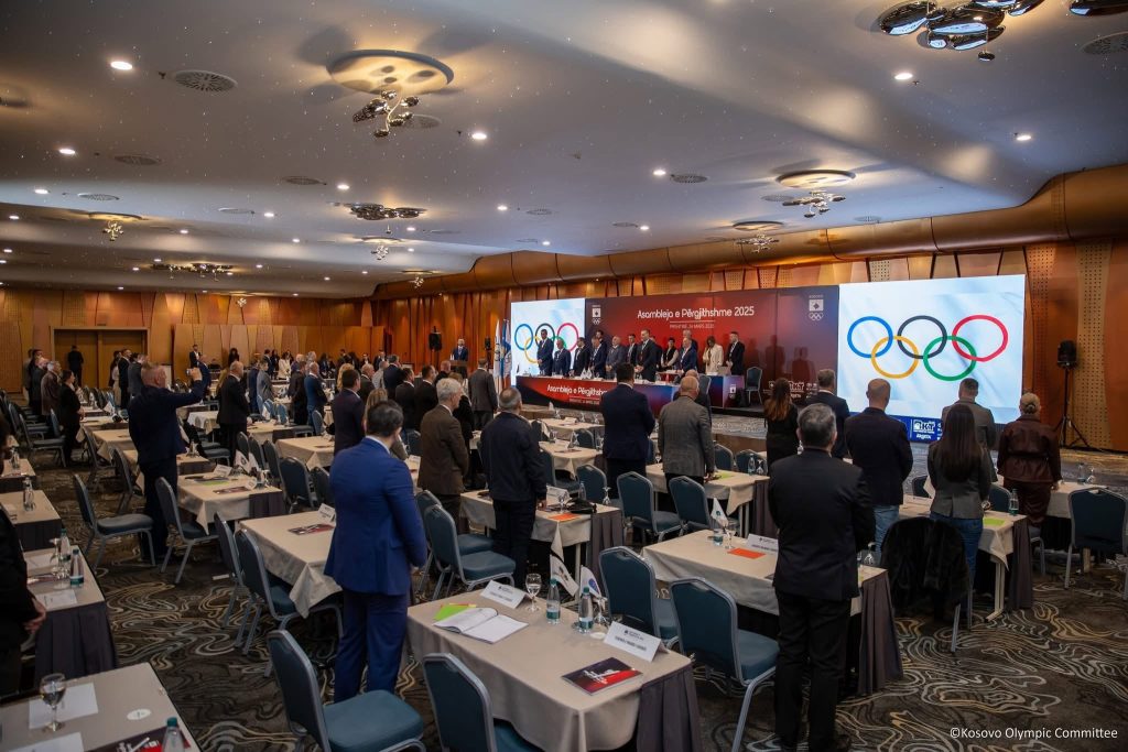 Salla e asamblesë me pjesëmarrës në këmbë dhe rrathët olimpikë në ekran.